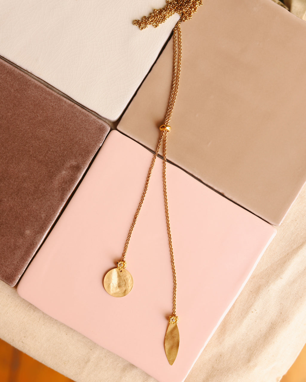 Colar up & down dourado com medalha martelada e folha, sobre azulejos rosa e neutros