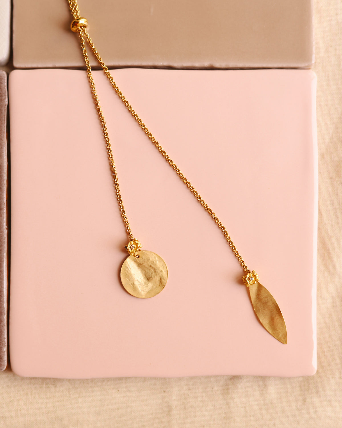 Colar up & down dourado com medalha martelada e folha, sobre azulejos rosa e neutros